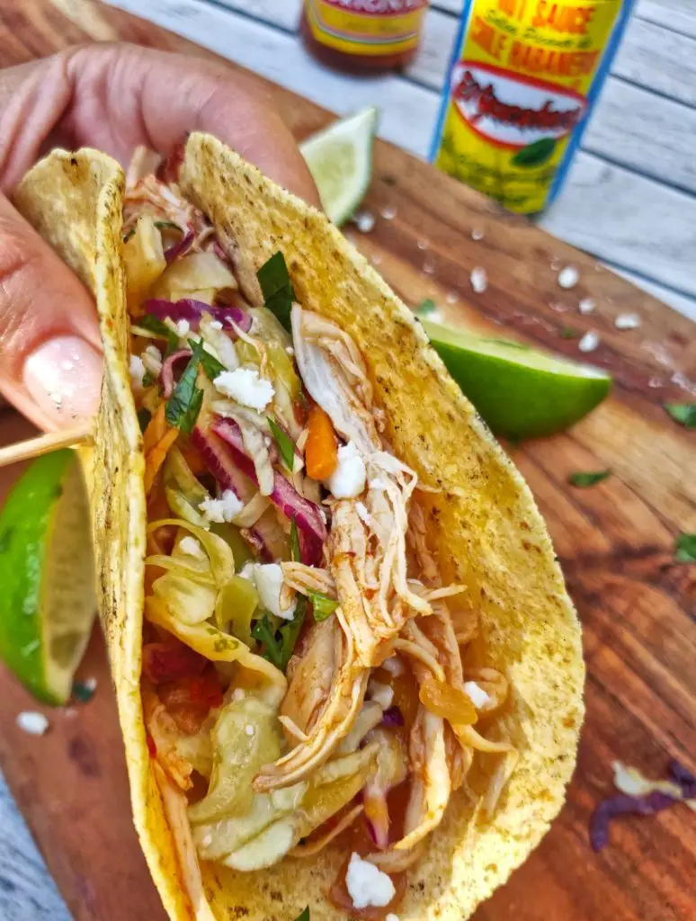 close up front image of hand holding chipotle chicken taco with mango lime salsa with hot sauce bottles in background and sliced limes