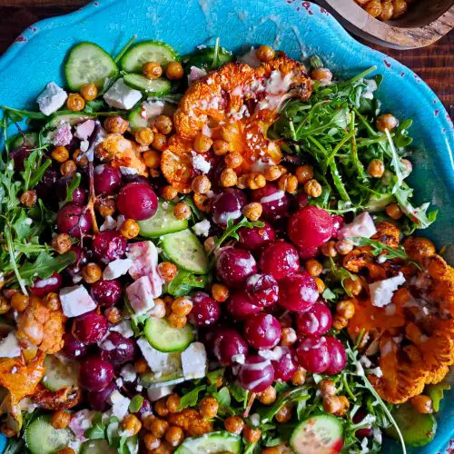 overhead image of middle eastern salad with harissa cauliflower roasted grapes and lemon tahini dressing next to wooden bowl of crispy chickpeas
