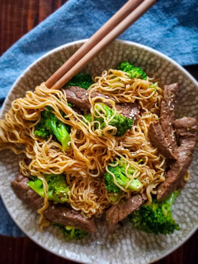 image of beef and broccoli ramen stir fry in bowl with chopsticks and blue linen napkin