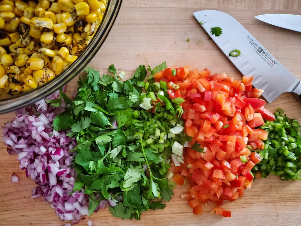 chopped onion, cilantro, red pepper, and jalapeno on cutting board with bowl of corn kernals 