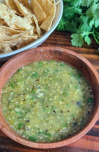 tomatillo salsa verde in small wooden bowl next to tortilla chips and sprig of cilantro