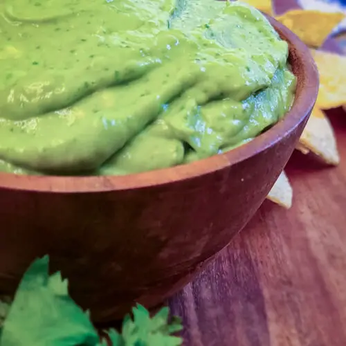image of a side view close up of a creamy whipped guacamole in a small wooden bowl with a handful of tortilla chips next to the guacamole
