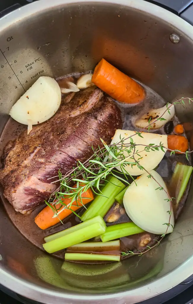 image of instant pot with seared beef in cooking liquid, add onion, garlic, celery, carrot, rosemary, and thyme