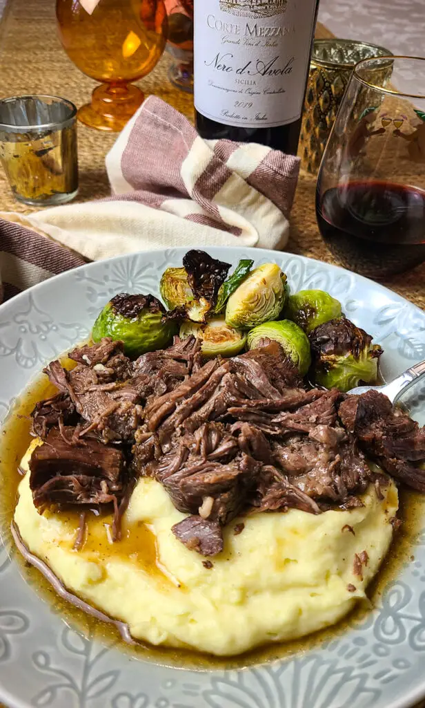 plate of shredded instant pot chuck roast with gravy over mashed potatoes with side of roasted brussels sprouts on dining table with red wine and gold votives. 