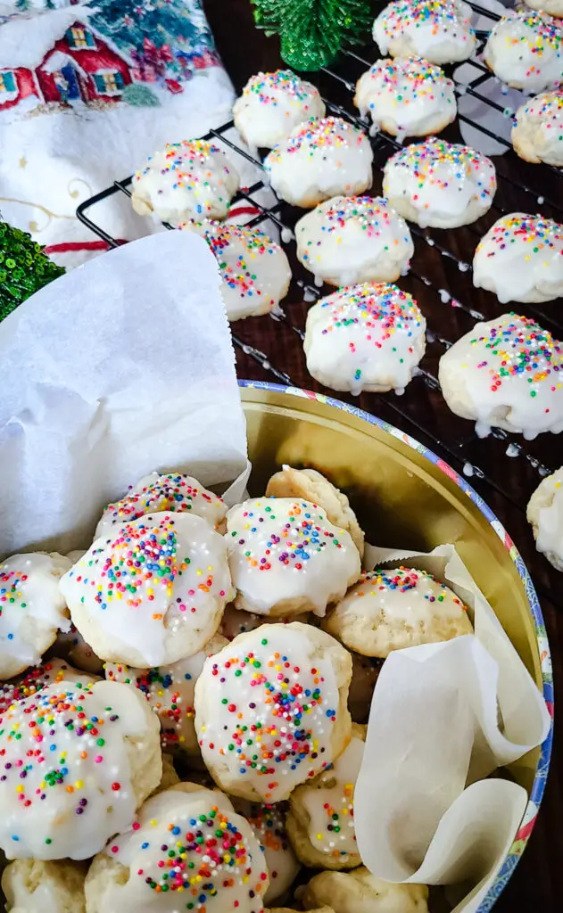 image of italian lemon ricotta cookies in a tin next to a more cookies on a wire rack with a christmas themed tea towel on the table