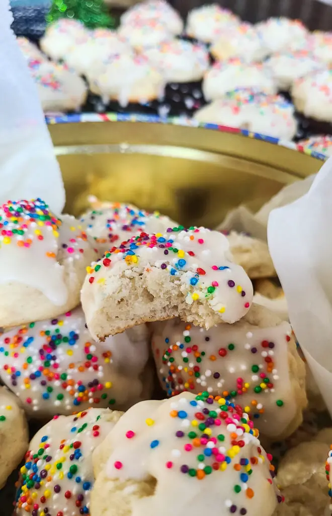 image of a bitten lemon ricotta sprinkle cookie resting a top of a cookie tin filled with whole cookies