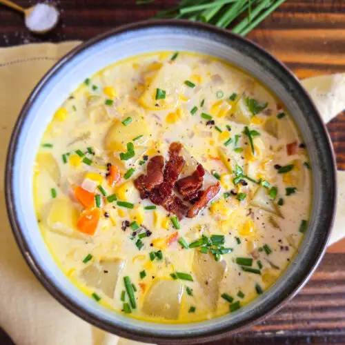 Close-up of a bowl of creamy fresh corn chowder with Yukon gold potatoes, crispy bacon, diced carrots, and chopped chives in a blue-rimmed bowl