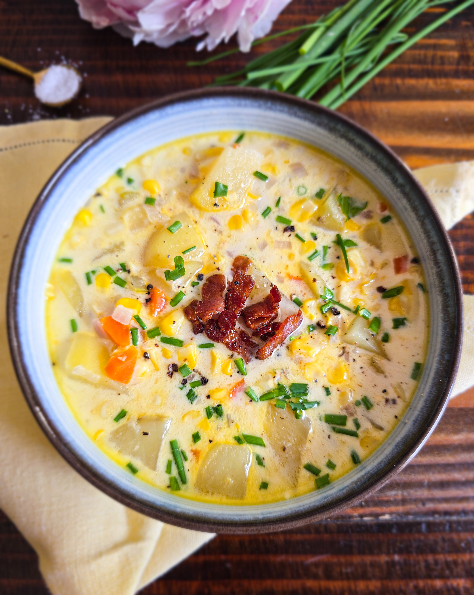 Close-up of a bowl of creamy fresh corn chowder with Yukon gold potatoes, crispy bacon, diced carrots, and chopped chives in a blue-rimmed bowl